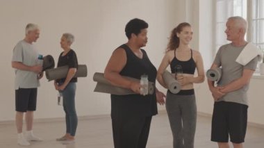 Kollarının altında yoga minderleri olan, geniş minimalist yoga stüdyosunun önünde genç bayan yoga hocasıyla konuşan spor giyimli çeşitli son sınıf öğrencilerinin orta boy fotoğrafları.