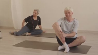 Beyaz saçlı, spor giyimli, kameraya gülümseyen, yoga minderinin üstünde oturan beyaz saçlı son sınıf öğrencisinin portresi. Olgun Asyalı kadın arka planda yoga yaparken.