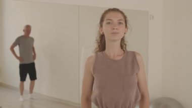 Geniş dans stüdyosundaki yaşlıları aynalarla eğitirken kameraya bakan beyaz kadın dans hocasının orta boy fotoğrafı.
