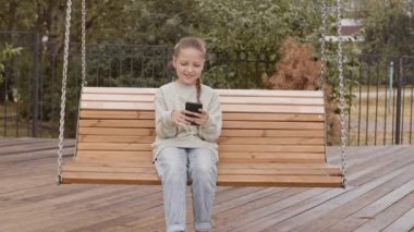10 yaşında, rahat giyimli, V işareti gösteren, akıllı telefondan selfie çeken, yaz mevsiminde oyun parkındaki ahşap salıncakta oturan beyaz bir kızın yavaşlığı.