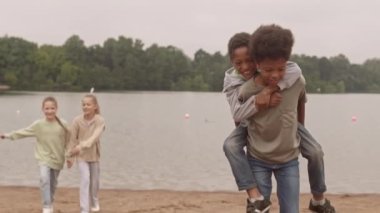 Yazı dışarıda göl kenarında geçiren çeşitli okul öğrencilerinin yavaşlığı. Küçük kardeşini omuzlarında taşıyan Afrikalı Amerikalı bir çocuk ve arka planda el ele koşan iki beyaz kız.