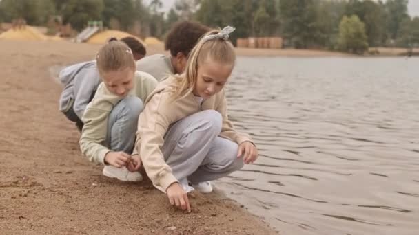 ゆっくりのかなり10歳の白人の女の子と彼女の多人種間の友人話している間に石を収集砂の上に座っている湖でビーチ — ストック動画