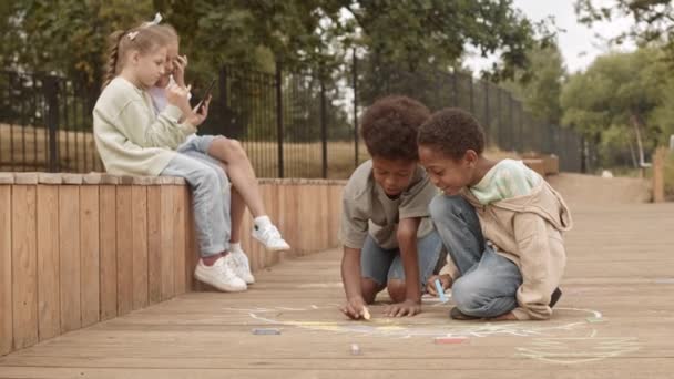 两个可爱的非洲裔美国男孩慢吞吞地在木板上画粉笔 而两个漂亮的白人女孩则在后面的智能手机上打滚 在公园里度过夏天 — 图库视频影像