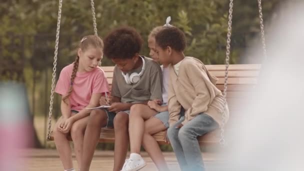 Τέσσερα Ευτυχισμένα Πολυεθνικά Παιδιά Κάθονται Ένα Παγκάκι Στην Παιδική Χαρά — Αρχείο Βίντεο