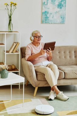 Modern yaşlı bir kadının dikey portresi. Kulaklıklı tablet kullanıyor. Akıllı ev iç dekorasyonunda koltukta oturuyor.