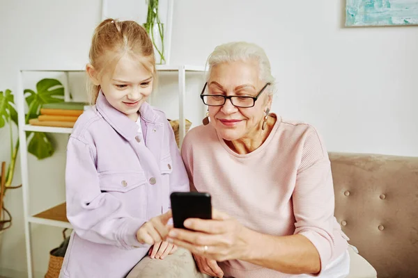 可愛いです女の子の肖像とともにおばあちゃんは居心地の良い家でスマートフォンの画面を見て — ストック写真