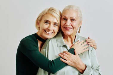 Stüdyoda sarılıp kameraya bakan yetişkin bir kızı olan yaşlı bir kadının samimi belden yukarı portresi.