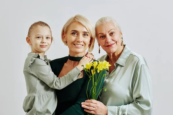 Ailede elinde çiçekler olan ve stüdyoda kameraya gülümseyen üç kadının minimum portresi.
