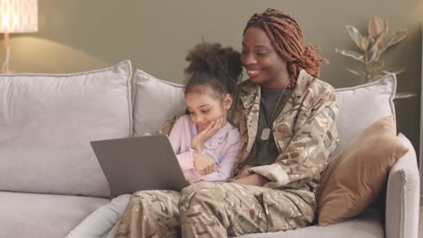 中遅いです幸せな若いです黒女性で軍制服と彼女の可愛いです6歳の娘使用してノートパソコン一緒に座っていますソファに自宅 — ストック動画