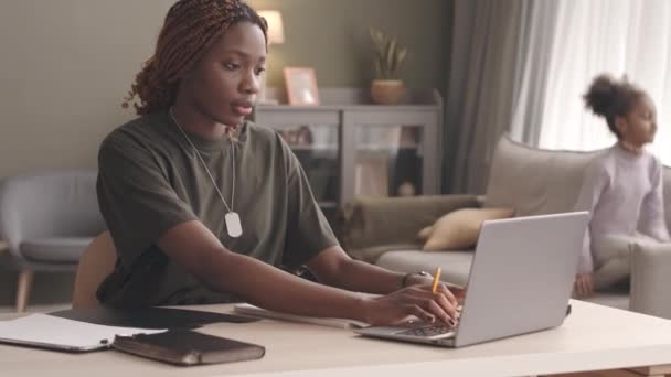 年轻的黑人女兵在家里用笔记本电脑干活 而她那嬉闹的小女儿在沙发上做背景时 动作有点慢 — 图库视频影像