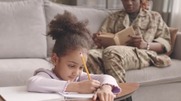 ゆっくりのかなりアフリカ系アメリカ人の女の子あります宿題でリビングルーム中に彼女のお母さんで軍制服読書本座っている間に柔らかい灰色のソファに背景 — ストック動画