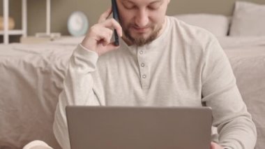 Nilüfer çiçekli genç beyaz iş adamının yavaş yavaş yatağında dizüstü bilgisayarla poz verip evden telefon görüşmesi yapması.