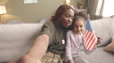 Ellerinde ABD bayrağı olan genç siyahi kadın asker ve küçük kızının el kamerasıyla selfie çekerken, oturma odasındaki yumuşak gri kanepede otururken çekilmiş resmi.