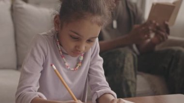 Askeri üniformalı babası arka planda koltukta oturmuş kitap okurken Afrikalı Amerikalı küçük bir kızın matematik ödevini yapmasıyla orta boy yakın çekim.
