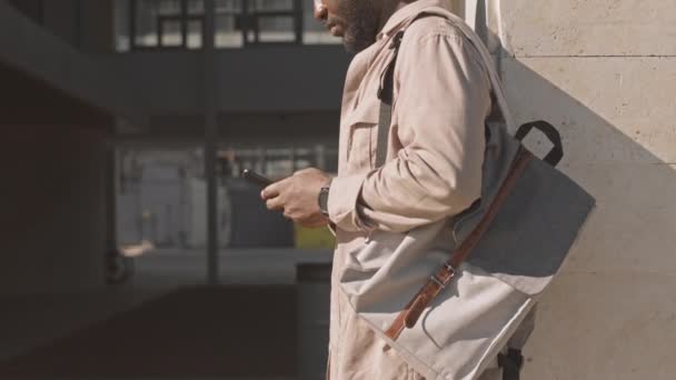 在阳光明媚的日子里 穿着米色衣服 背着手提电脑 背着纺织品背包 站在室外打电话的年轻黑人男子放慢了脚步 — 图库视频影像
