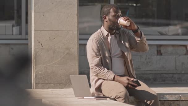 阳光明媚的日子里 穿着米色衣服的中等速度黑人年轻人坐在户外的长椅上 一边喝着外卖杯咖啡 一边打电话 — 图库视频影像
