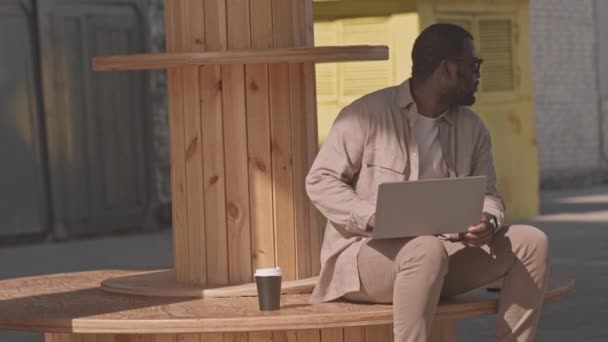 在阳光灿烂的日子里 穿着米色衣服 头戴深色墨镜的美国黑人商人一边在户外笔记本电脑上工作 一边慢慢地喝着外卖杯中的咖啡 — 图库视频影像