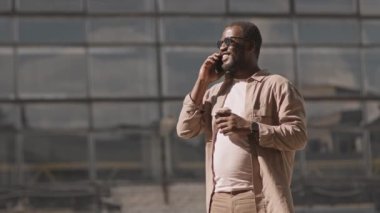 Beyaz elbiseli ve koyu güneş gözlüklü başarılı Afro-Amerikan işadamlarının orta yavaşlığı elinde hazır kahve bardağıyla akıllı telefondan konuşuyor, cam ofis binasına karşı ayakta duruyor.