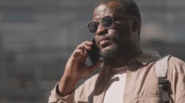 Koyu güneş gözlüklü ve bej rengi günlük giysili, elinde hazır kahveyle güneşli bir günde açık havada duran akıllı telefondan çağrı yapan Afro-Amerikalı genç bir adamı yavaşça yukarı kaldır.