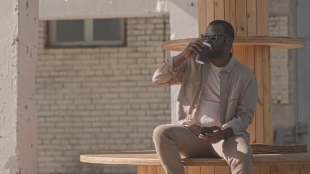 穿着米黄色便服和深色衣服的非洲裔美国商人慢吞吞地喝着外卖杯中的咖啡 在阳光灿烂的日子里坐在户外用智能手机聊天 — 图库视频影像