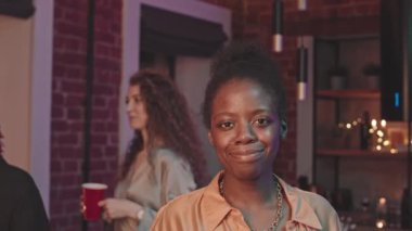 Ev partisinde kameraya gülümseyen genç, güzel siyahi bir kadının belden aşağı portresini kaldırırken arka planda elinde plastik bardaklarla sohbet eden kadın arkadaşları...