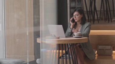 Kafenin penceresinde dizüstü bilgisayarıyla oturan ve akıllı telefondan konuşan esmer bir iş kadını.