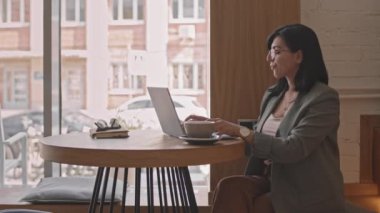 Zeki günlük kıyafetleri ve gözlükleri olan, kahve içen ve rahat bir kafede oturup dizüstü bilgisayarda çalışan çekici, çift ırklı genç iş kadınının orta derecede yavaşlığı.