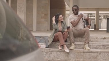 Yavaş yavaş, akıllı telefondaki siyah güneş gözlüklü Afrikalı Amerikalı erkek arkadaşına bir şey gösteren esmer kadın yazın açık havada merdivenlerde oturuyor.
