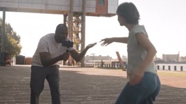 Genç siyahi bir adamın kamerasıyla sokak dansı yapan beyaz bir kadının orta yavaşlığı onu neşelendiriyor, güneşli bir günde açık havada video çekiyor.