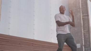 Beyaz tişörtlü, neşeli, genç Afrikalı Amerikalı adam gün ışığında dışarıda dans ediyor.