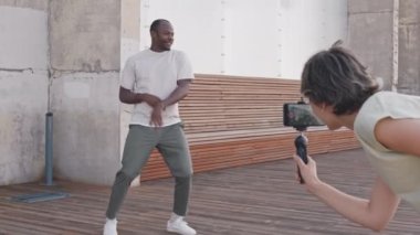 Güneşli bir günde açık havada dans eden enerjik genç siyahi adamın tam boy çekimi. Bu sırada bayan arkadaşı onu akıllı telefonla kaydediyor ve sosyal ağlar için video dersi veriyor.