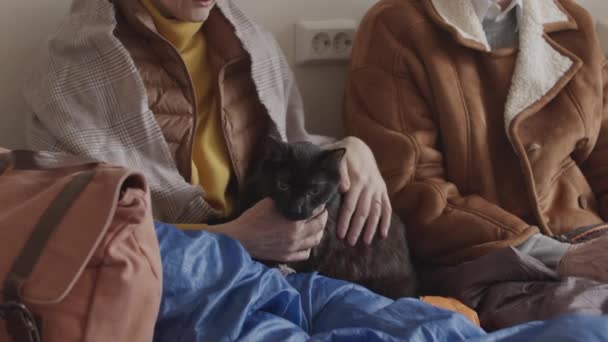 屋内の空き部屋で他の難民と一緒に座っている古い暖かい服を着て成熟した難民の女性の作物の減速ホームレスの黒猫をペット — ストック動画