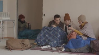 Eski giysiler ve uyku tulumlarıyla birlikte sığınakta oturmuş cep telefonlarından internette gördükleri politik haberleri tartışan genç ve yetişkin mültecilerden oluşan yavaş bir grup.