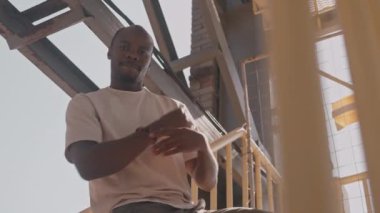 Sıcak güneşli bir günde inşaat alanının sarı merdivenlerinde hip hop dansı yaparken enerjik siyahi bir adamın kameraya baktığı düşük açılı çekim.