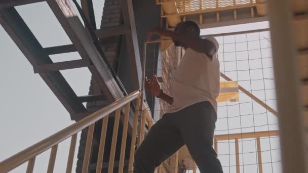 站在工地黄色楼梯上 站在阳光下 身披白色T恤的非洲裔年轻男子的低角度慢动作 动作塑性变形 — 图库视频影像