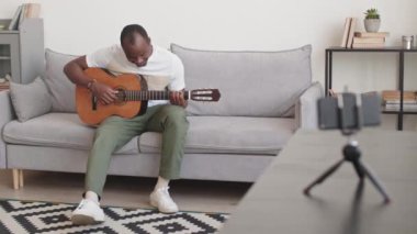 Akustik gitarla kanepede otururken kameraya el sallayıp gülümseyen, akıllı telefondan video gitar dersi alan neşeli siyahi bir adamın istatistik çekimi.