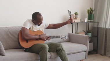 Elinde akıllı telefon olan ve akustik gitar çalarken videosu olan siyah bir adamın evdeki kanepede oturmasının yavaşlaması.