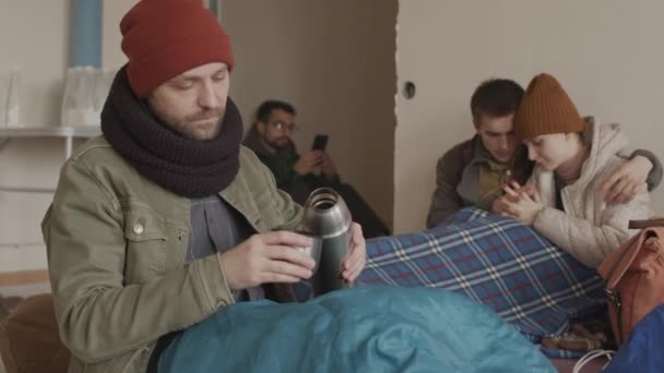 寒い避難所に一緒に滞在し 古い毛布や寝袋で暖まる 魔法瓶から熱いお茶を注ぎ 他の難民にそれを提供し 髭の難民の男の遅い — ストック動画