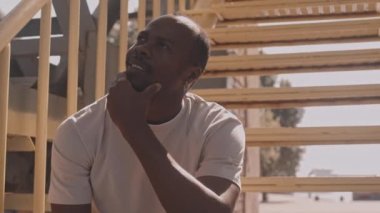 Sıcak, güneşli bir günde inşaat alanının sarı merdivenlerinde otururken gökyüzüne bakıp rap yapan dalgın genç bir zenci.