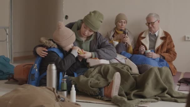 他の難民と一緒に寒い避難所に滞在しながら 彼女の7歳の息子にサンドイッチを食べさせる若い白人難民女性のスローモ — ストック動画