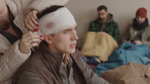 負傷した頭部を持つ若い難民男性の中程度の減速は 冷たい亡命の小さなグループに一緒に住んで 女性難民によって包帯を頭を持って 最初の医療援助を与えられました — ストック動画