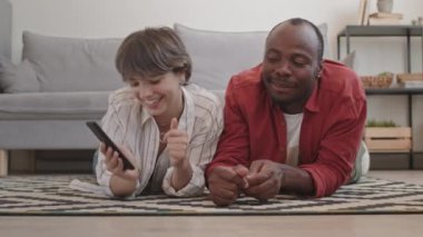 Akıllı telefondan komik dans videoları seyreden ve sürekli hareketler yapan, oturma odasında karnının üzerinde uzanan mutlu ve çeşitli çiftlerin yavaş yavaş gülüşleri.