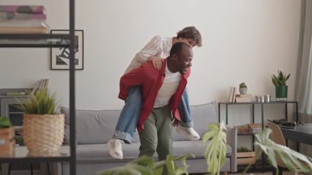 一个快乐的黑人小伙子背着他那漂亮的白人女朋友在客厅里打转 一边笑一边笑 — 图库视频影像