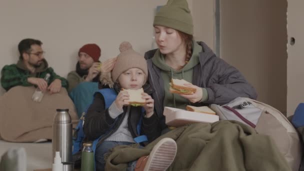 白种人年轻难民妇女和她穿着保暖衣服的小儿子慢吞吞地吃三明治 而与其他难民一起坐在冰冷的庇护所的地板上 — 图库视频影像