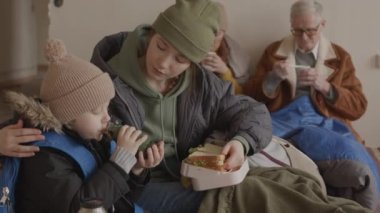 Küçük oğlunu sandviçle beslerken diğer mültecilerle birlikte soğuk ve rahatsız bir tımarhanede kalan, arka planda duvarda oturan olgun çift de dahil olmak üzere, umursamaz, genç, beyaz bir kadının ağırlığı.