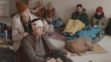 Ağır yaralı kafayla sığınma evinin soğuk zemininde oturan ve diğer evsizlerle birlikte yaşayan mültecilere ilk yardımı yapan genç kadın.