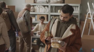 Sakallı mültecinin sakallı portresi ellerinde yiyecek ve su malzemeleriyle ne yazık ki kameraya bakarken içeride gönüllülerin sığınmacılara yardım ve yataklık ettiği yerde duruyor.