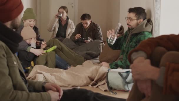 成群结队的难民和移徙者彼此分享食物和水供应 同时穿着旧衣服和睡袋坐在冰冷的庇护所地板上 — 图库视频影像