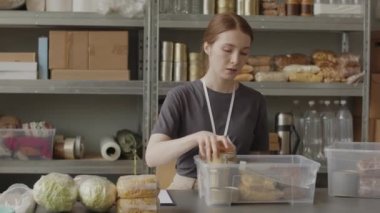 Beyaz kadın gönüllülerin orta yavaş portresi yerel gıda bankasında çalışan plastik konteynıra temel gıda erzaklarını koyuyor.