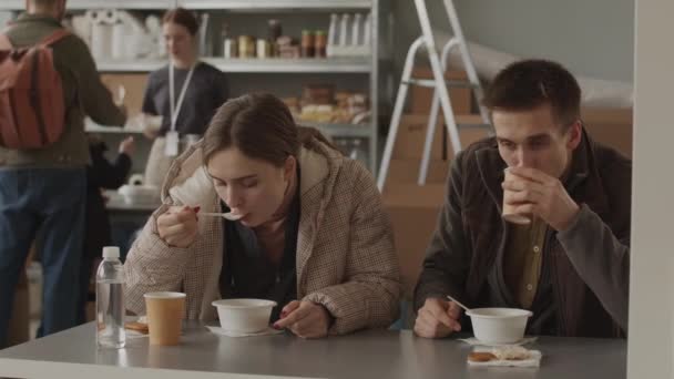 地元のフードバンクのテーブルに座っている使い捨て食器から熱いスープを食べる難民の若い白人のカップルの中低速背景に彼らの部分のキューに立っている人々 — ストック動画
